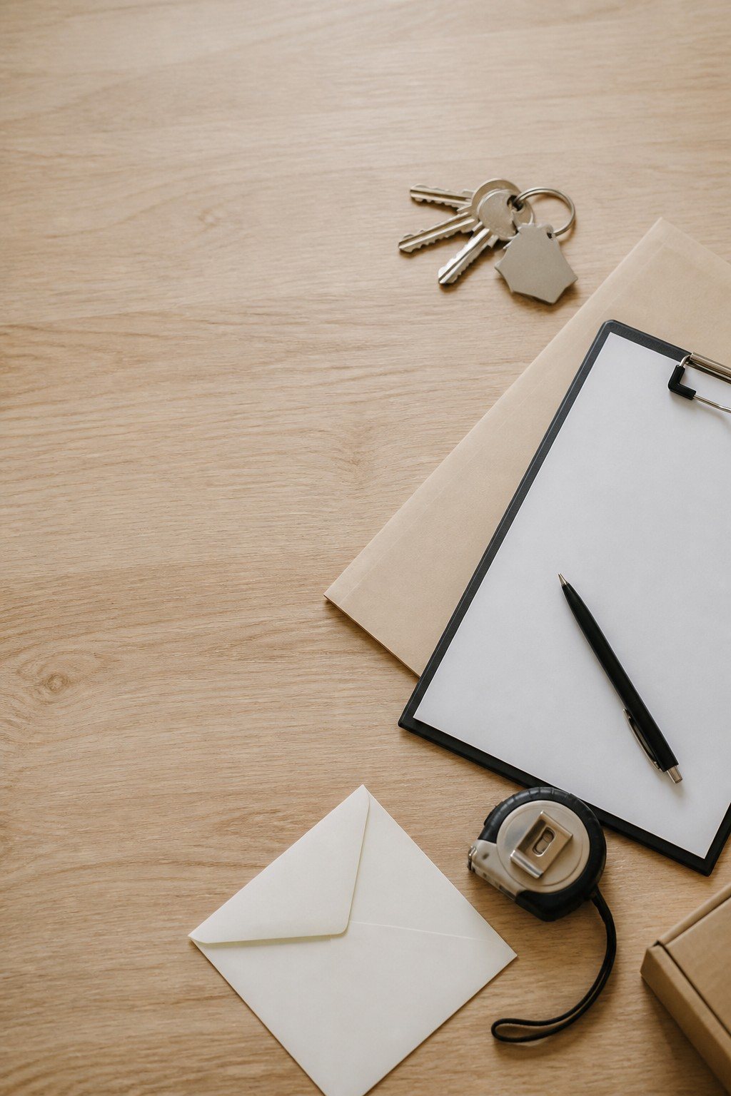 A clipboard, pen, envelope, and set of keys arranged on a wood desk